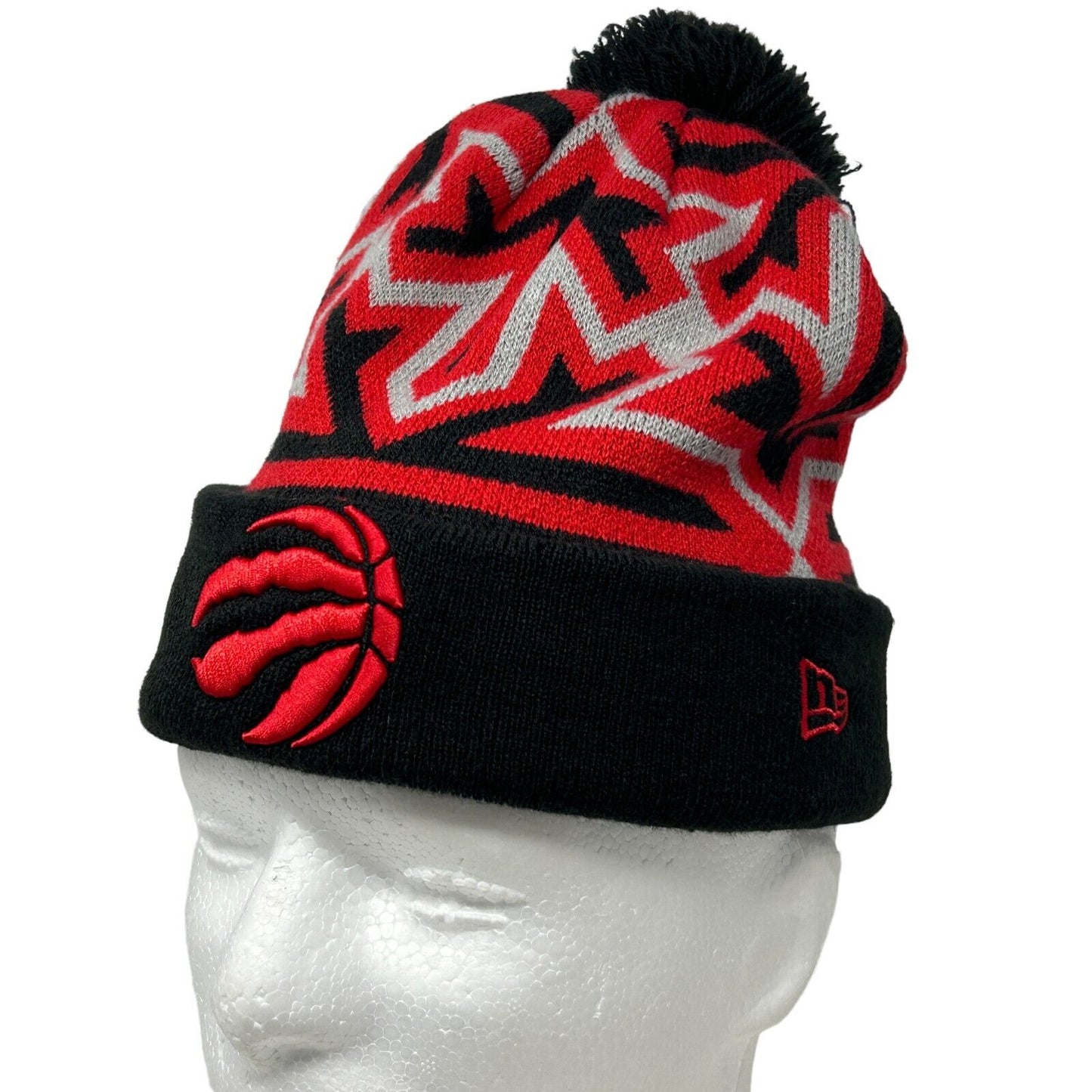 Red and black New Era Toronto Raptors beanie with a logo and pom-pom on top, front view detail.